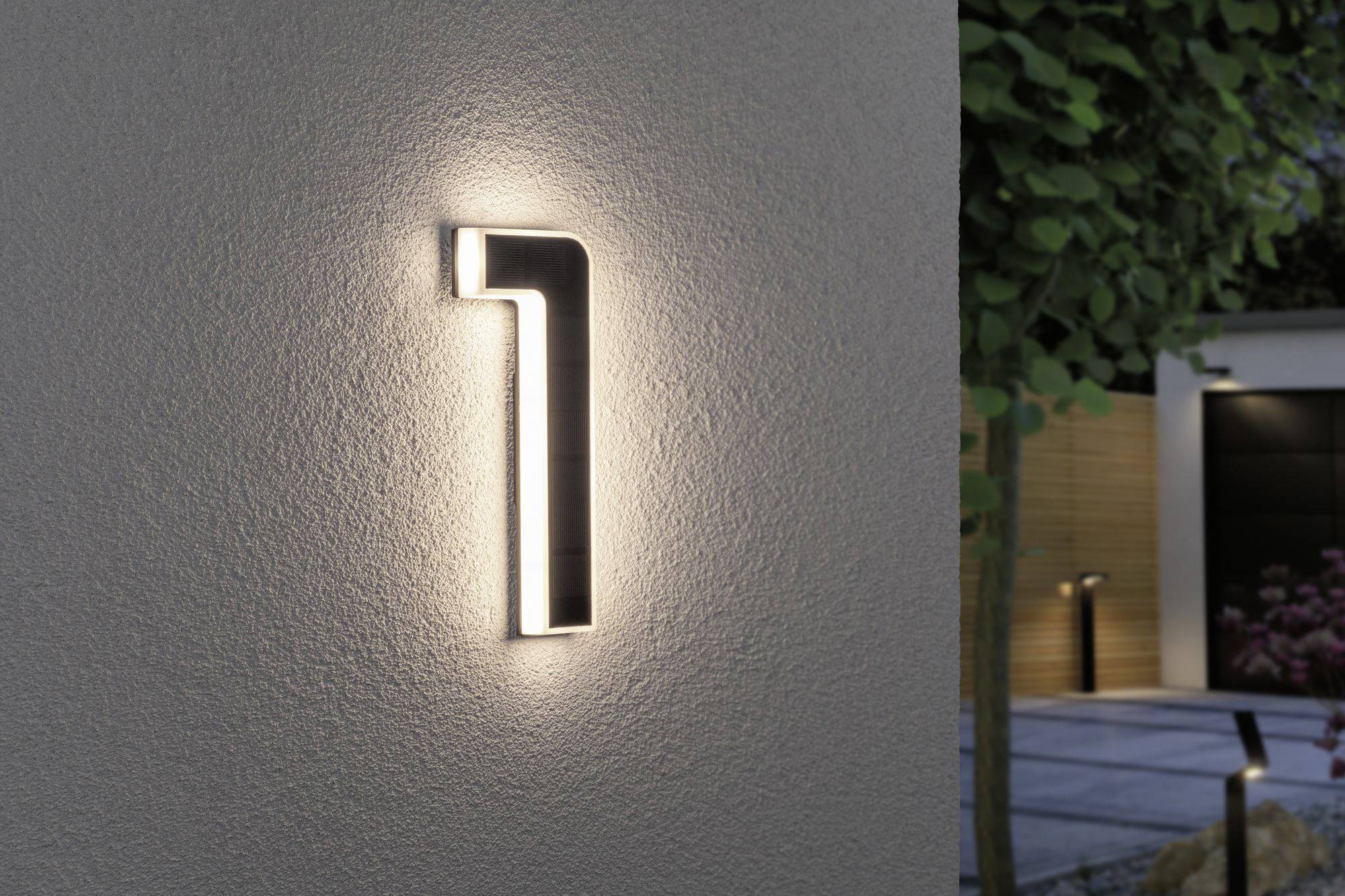 Un numéro de maison '7' illuminé sur un mur blanc, entouré de plantes vertes et d'un chemin pavé en arrière-plan.