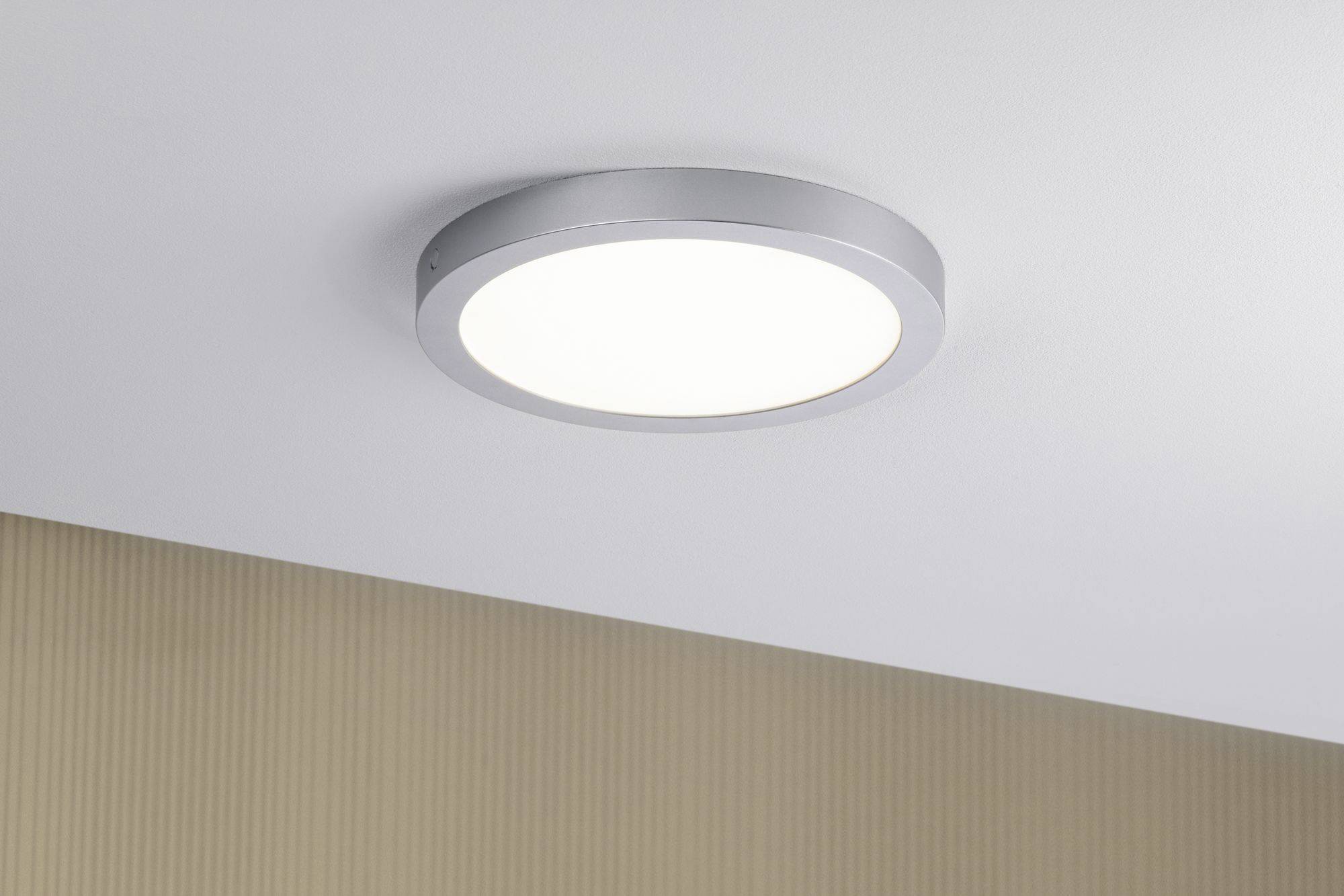 Lampe de plafond ronde avec cadre métallique illuminant un plafond blanc, un mur beige visible en dessous.