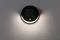 Une lampe solaire ronde accrochée à un mur gris éclaire en diffusant sa lumière vers le haut et vers le bas.