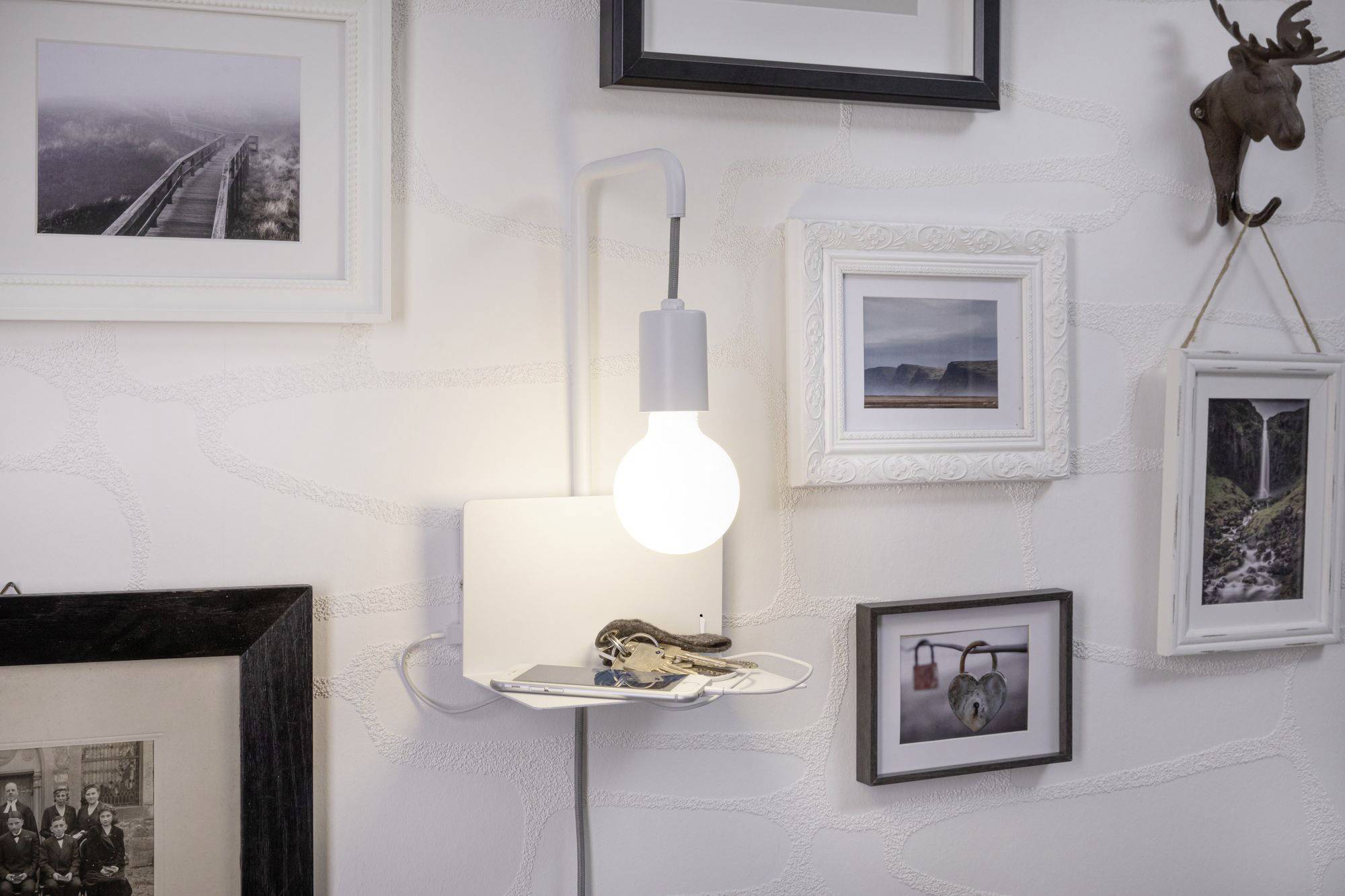 Plusieurs photos encadrées accrochées au mur, un abat-jour mural lumineux, en dessous une petite étagère avec des clés et des lunettes.