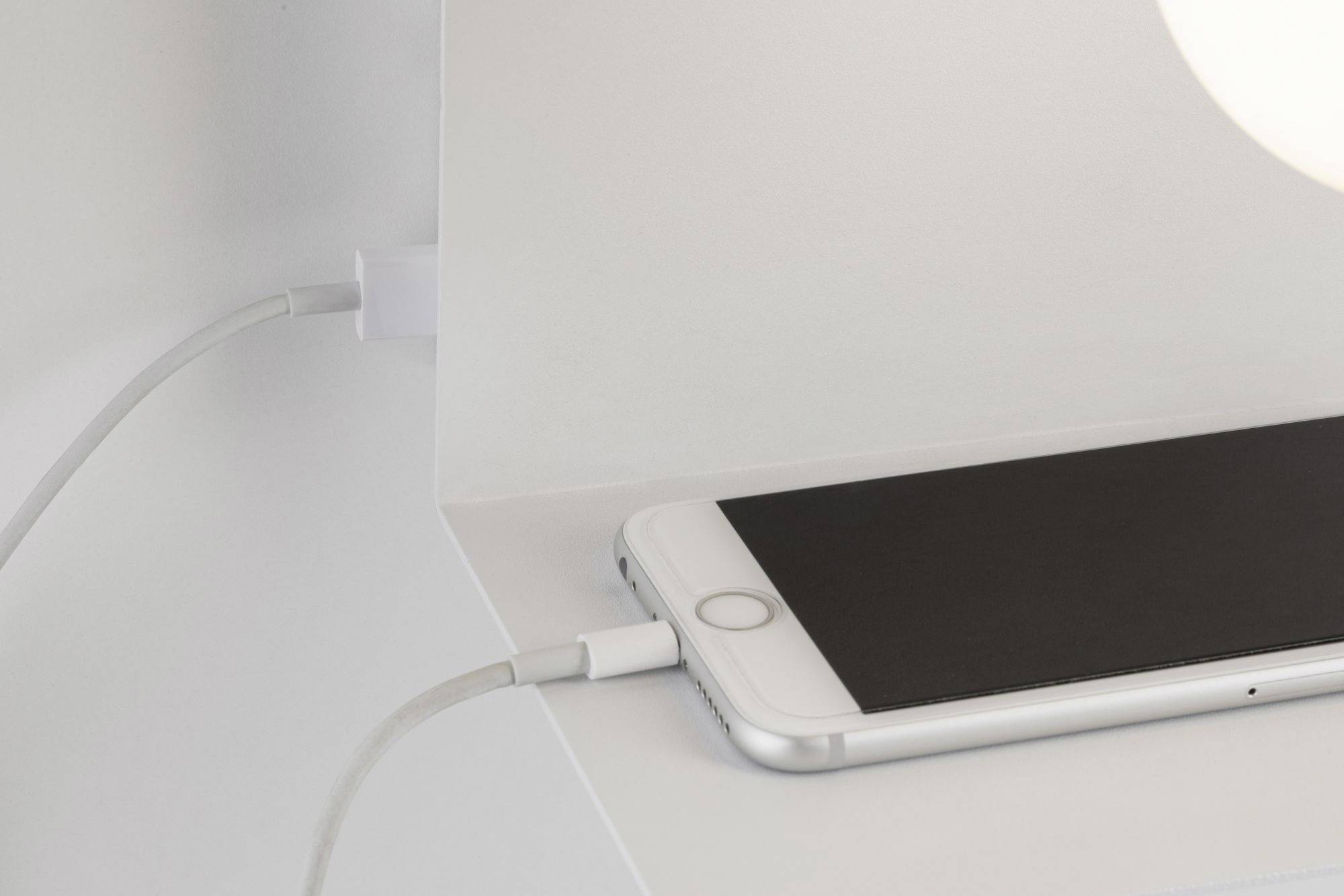Un smartphone est chargé à droite par un câble de charge. L'appareil est posé sur une surface blanche avec un fond blanc.