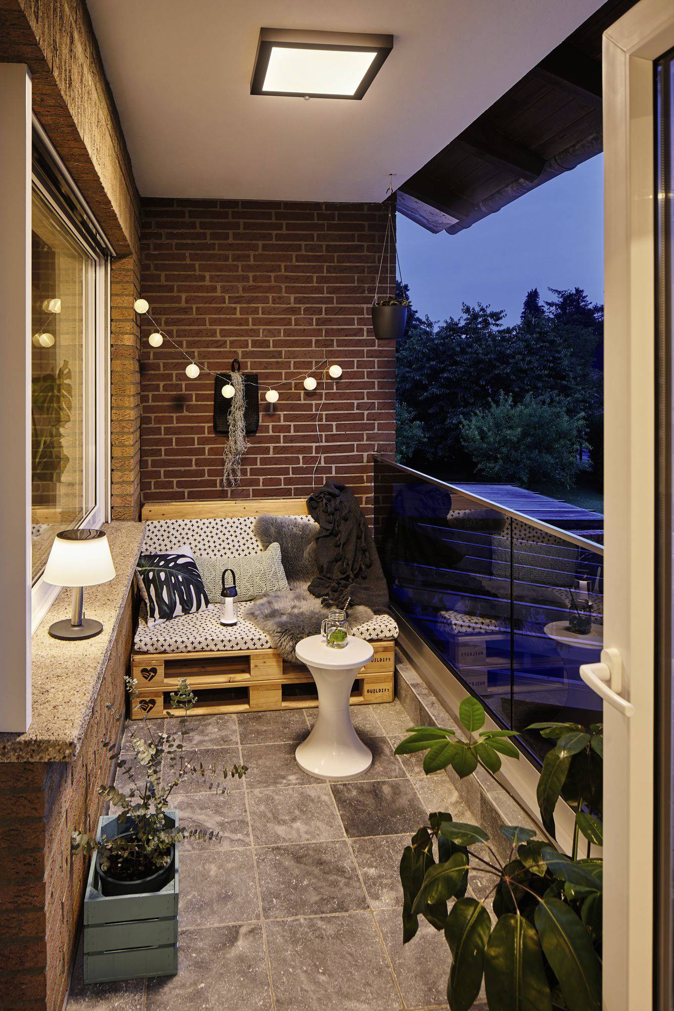 Petit balcon avec un coin salon et une plante. L'ambiance du soir, une guirlande lumineuse et une lampe de table créent un éclairage chaleureux et accueillant.