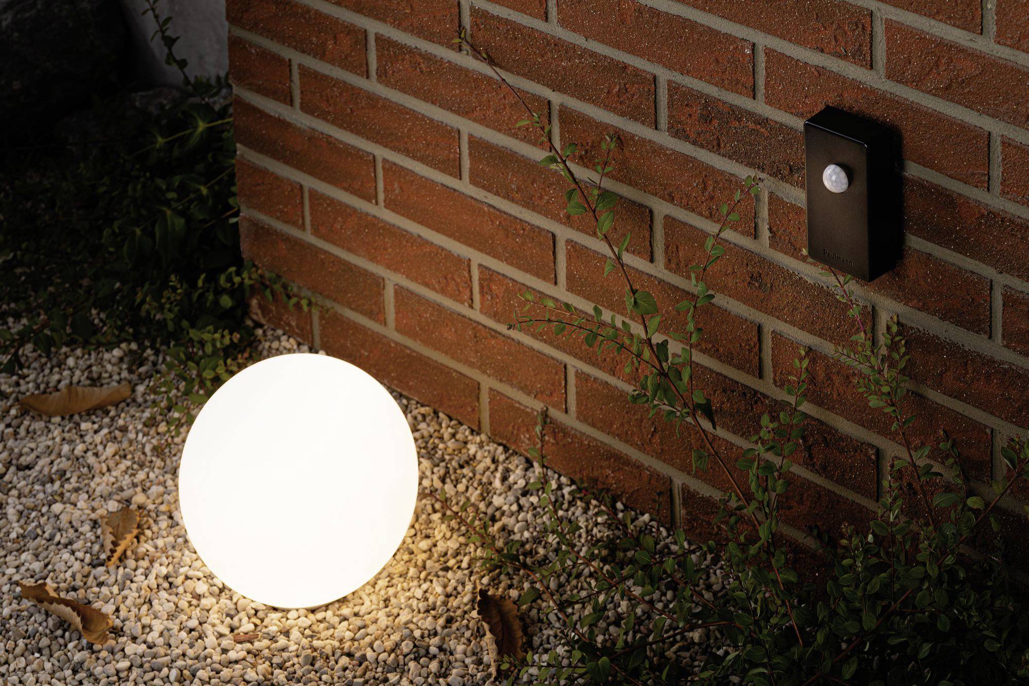 Une lampe sphérique lumineuse est posée sur du gravier devant un mur de briques, à côté duquel est fixé un détecteur de mouvement.