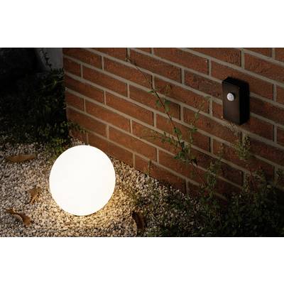 Une lampe sphérique lumineuse est posée sur du gravier devant un mur de briques, à côté duquel est fixé un détecteur de mouvement.