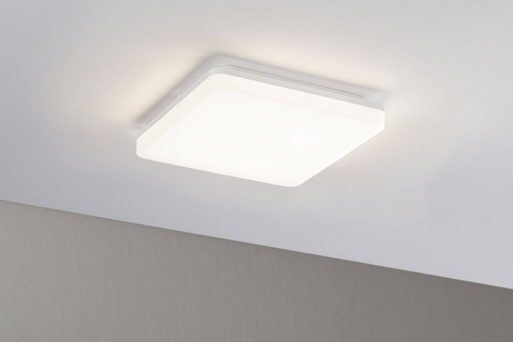 Un plafonnier carré à cadre blanc éclaire uniformément une pièce. Le plafond est clair, le mur en dessous est gris.