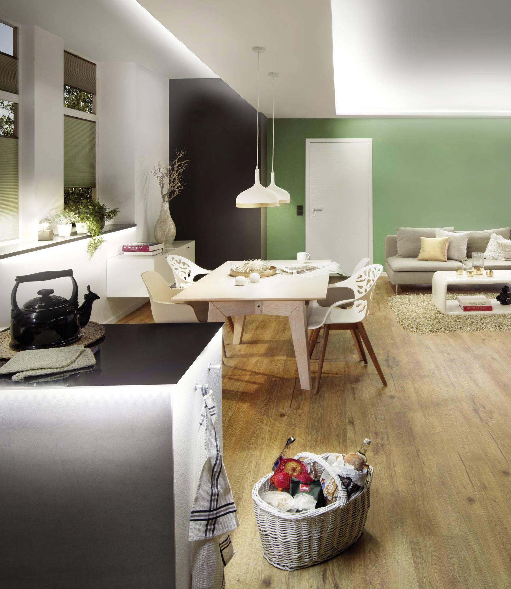 Cuisine ouverte et salon avec sol en bois. Murs clairs, table à manger avec chaises, canapé, lampes et éléments décoratifs. Panier avec courses posé au sol.