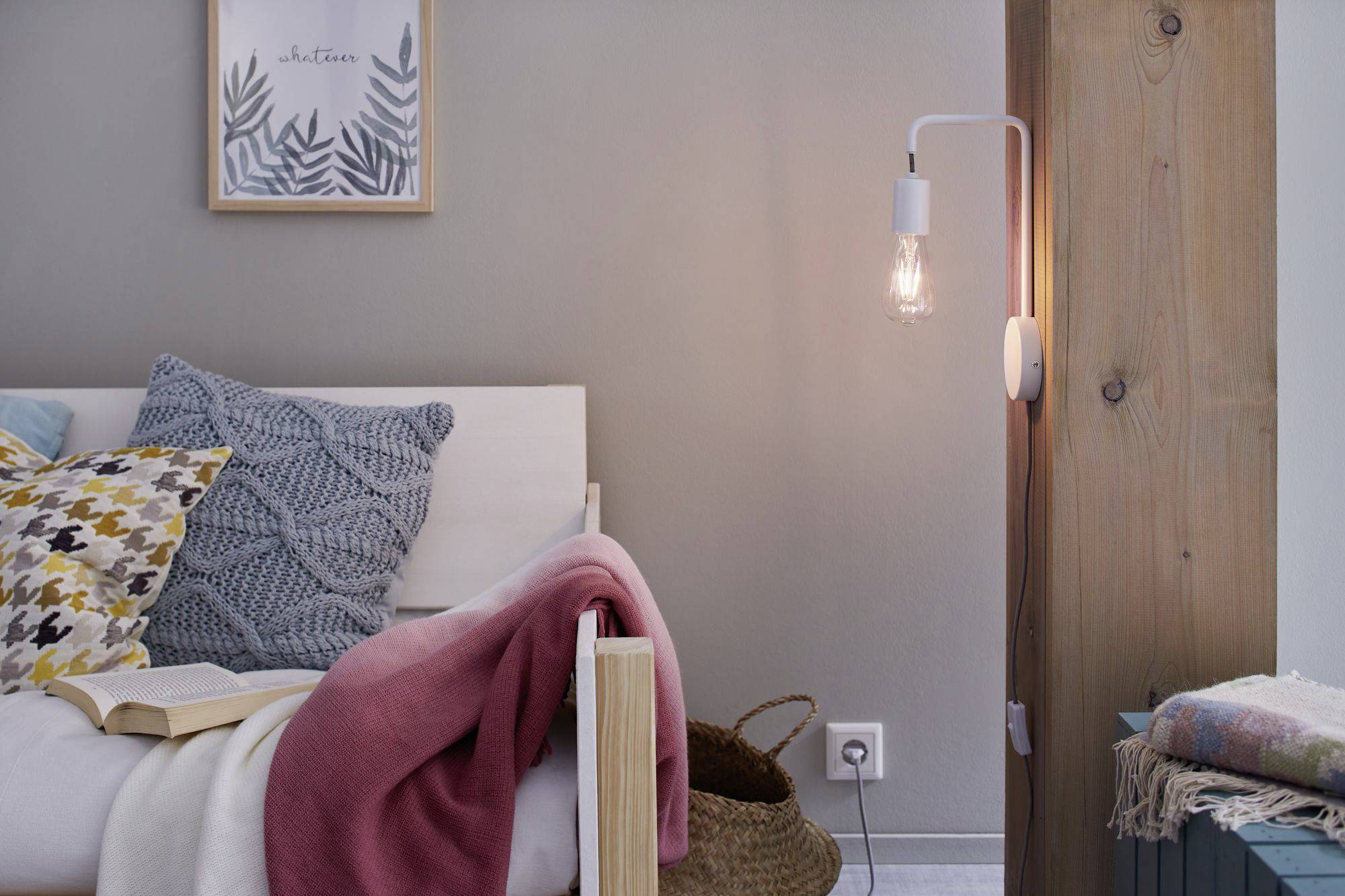 Une chambre douillette avec un lit rempli de coussins colorés, une couverture rose et un panier en osier à côté. Une lampe éclaire la pièce.