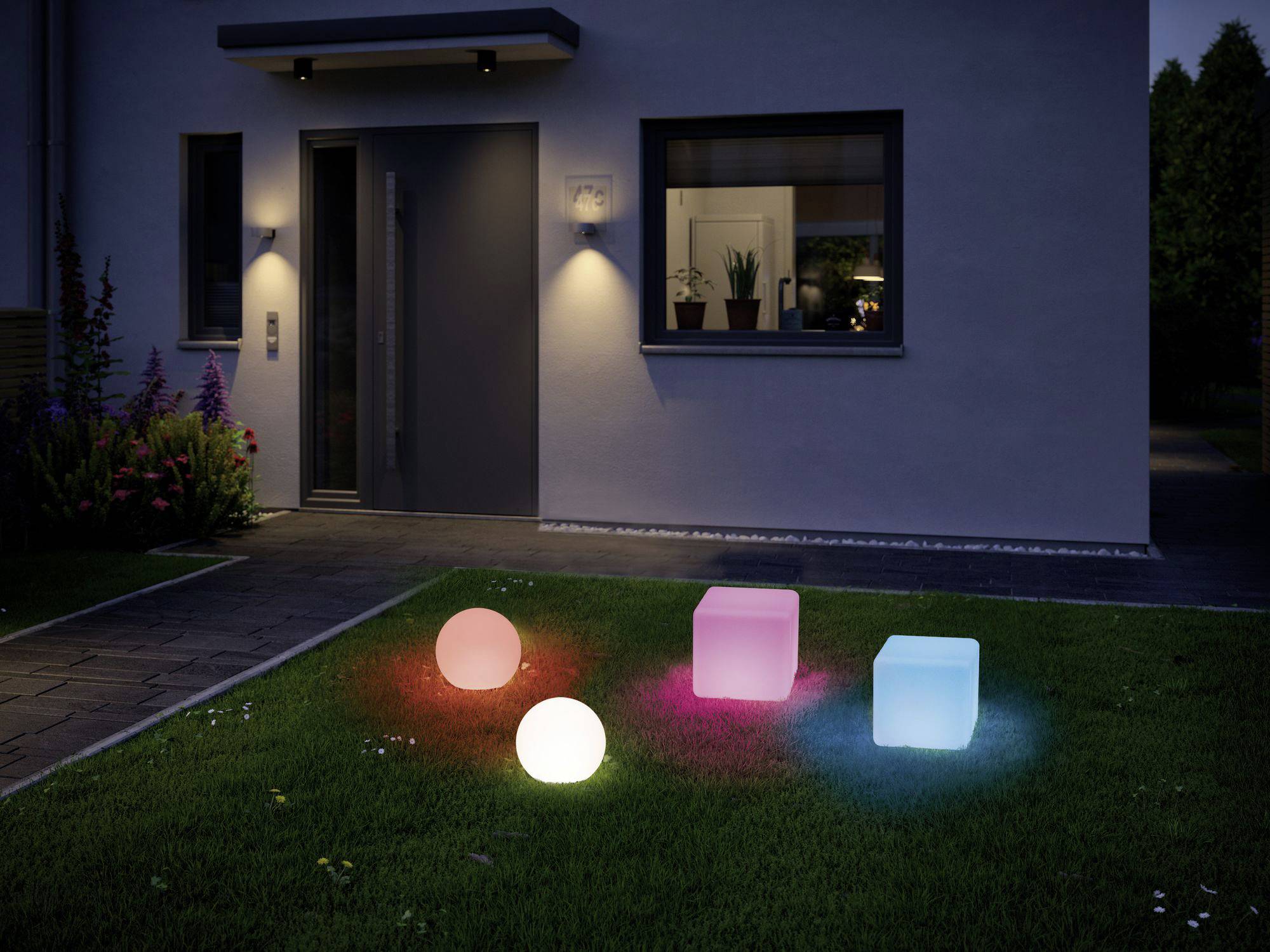 Jardin éclairé la nuit ; deux sphères et deux cubes de différentes couleurs rayonnent sur une pelouse devant une maison.