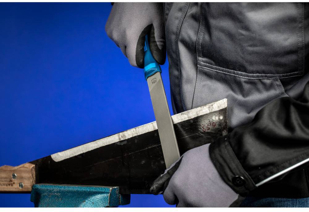 Une personne en tenue de travail et portant des gants affûte la lame d'un outil avec une lime sur un fond bleu.