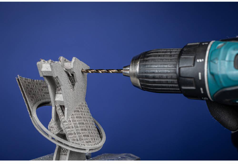 Un perceuse à batterie fore un objet en impression 3D gris aux formes complexes sur un fond bleu.