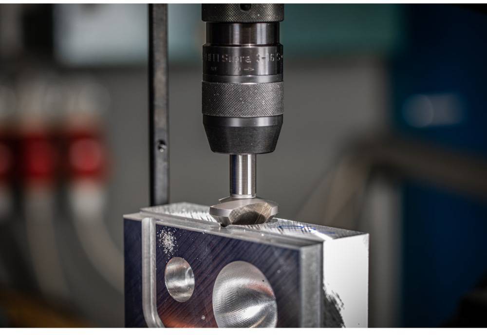 Une perceuse usine une pièce métallique avec des découpes précises. Des composants de machine rouges et bleus sont visibles en arrière-plan.