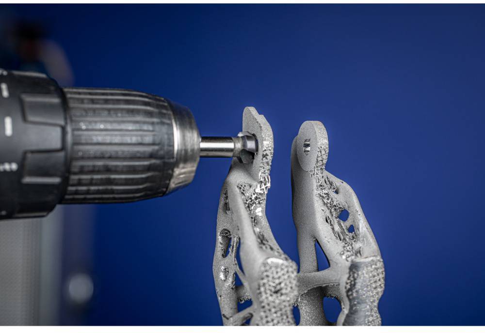 Une perceuse traite une pièce métallique complexe, imprimée en 3D, sur un fond bleu, illustrant un travail artisanal précis.