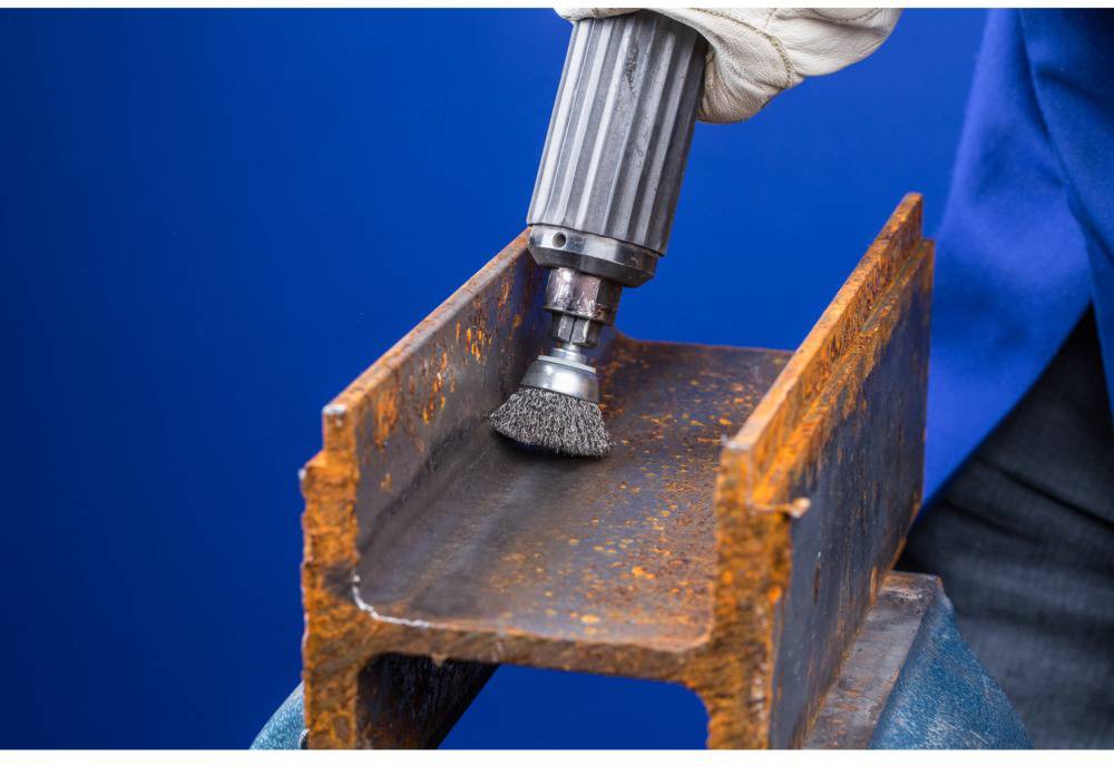 Une personne retire de la rouille d'un support métallique à l'aide d'une brosse métallique. Le support métallique est fortement corrodé.