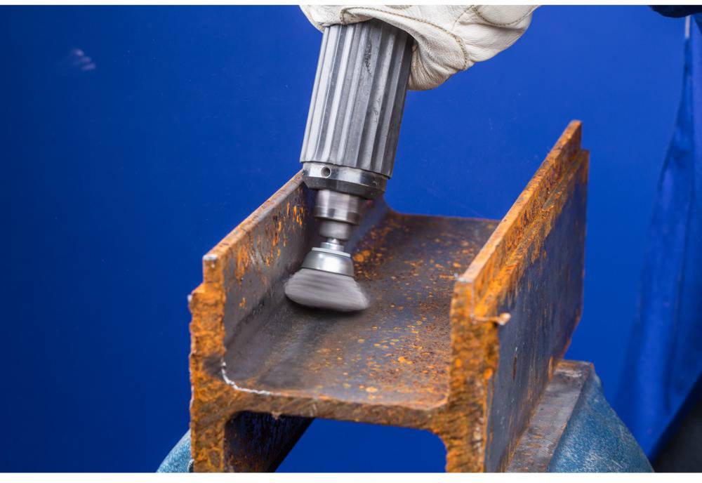 Une main tient un outil électrique avec un embout brosse, qui enlève de la rouille d'un support métallique. L'arrière-plan est bleu.