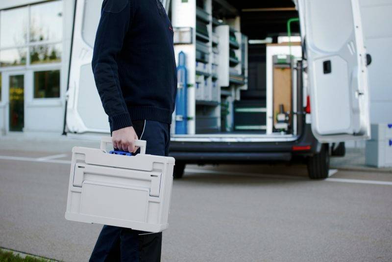 Une personne transporte une boîte à outils vers un van ouvert avec des étagères de rangement organisées, suggérant une préparation pour une tâche de service ou de réparation.