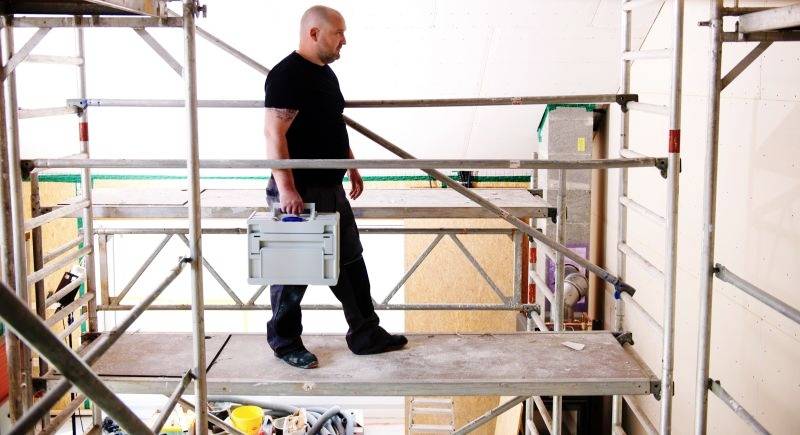 Une personne marche sur une plateforme d'échafaudage, transportant une boîte à outils. Le contexte semble être un chantier de construction ou de rénovation intérieur.