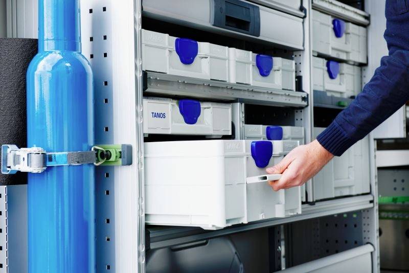 Une personne ouvre un tiroir dans une armoire de rangement remplie de contenants bleus et blancs bien organisés, à côté d'une bouteille de gaz bleue sécurisée.