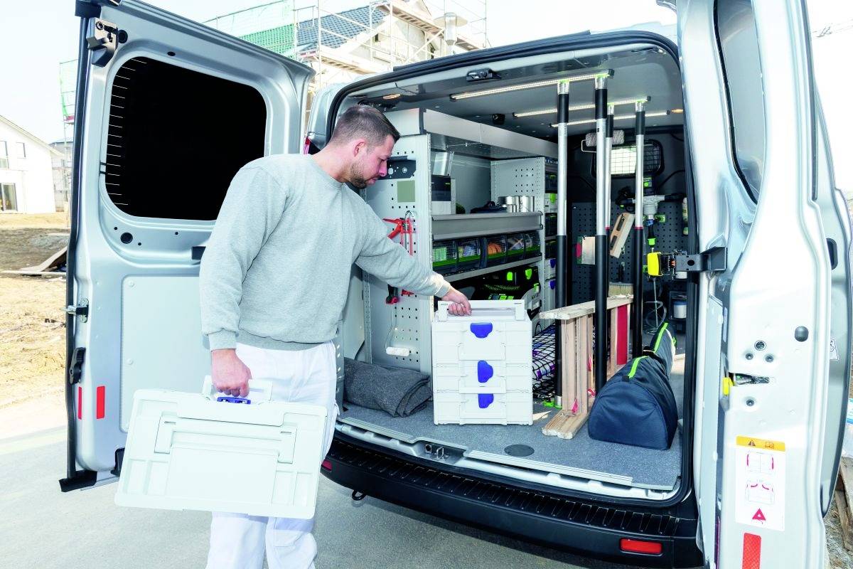 Une personne se tient à côté d'un fourgon ouvert rempli d'outils et d'équipements soigneusement rangés, en tendant la main vers une boîte à outils, ce qui suggère des travaux de réparation ou d'installation.