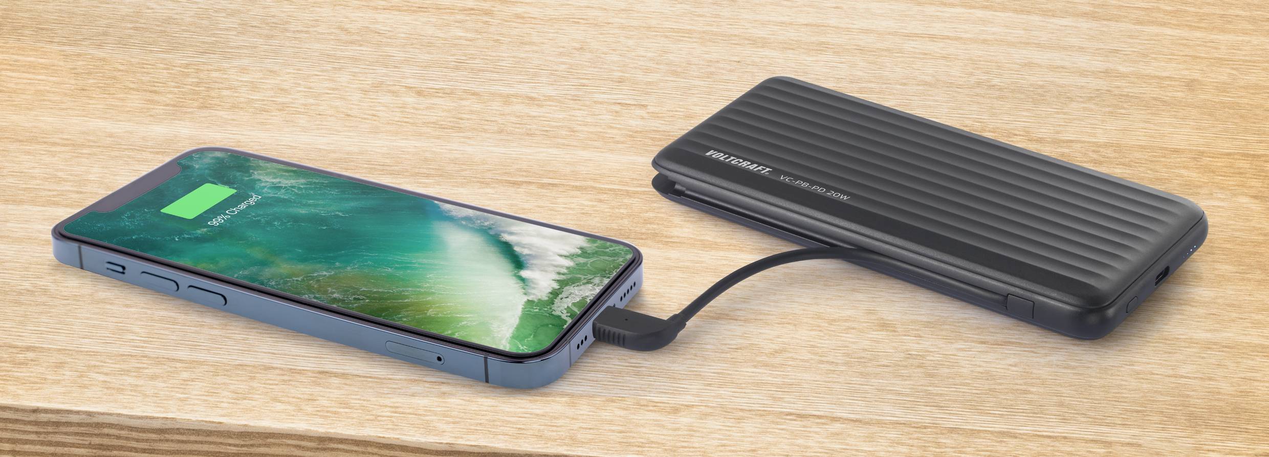 Un smartphone est en charge avec une batterie externe. L'écran affiche un symbole de batterie vert avec un indicateur de charge. Surface en bois en arrière-plan.
