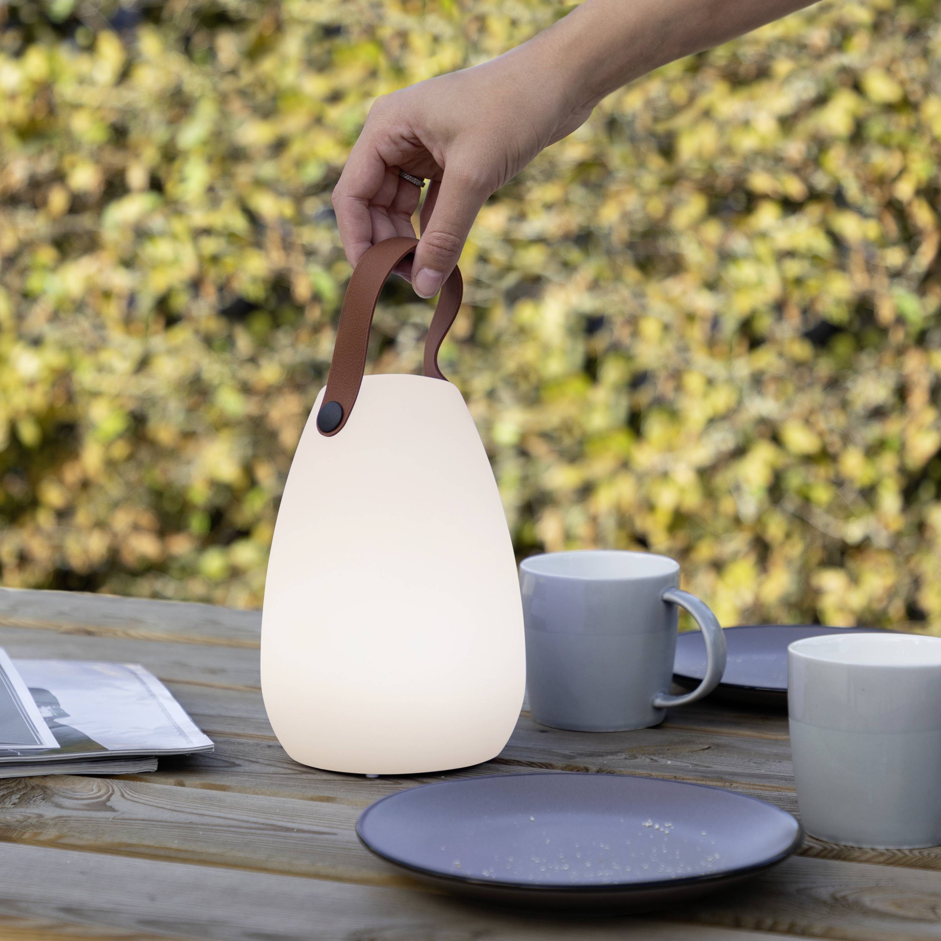 Une personne soulève une lampe portable avec une poignée en cuir d'une table en bois, sur laquelle sont posées des tasses et des assiettes, à l'extérieur.