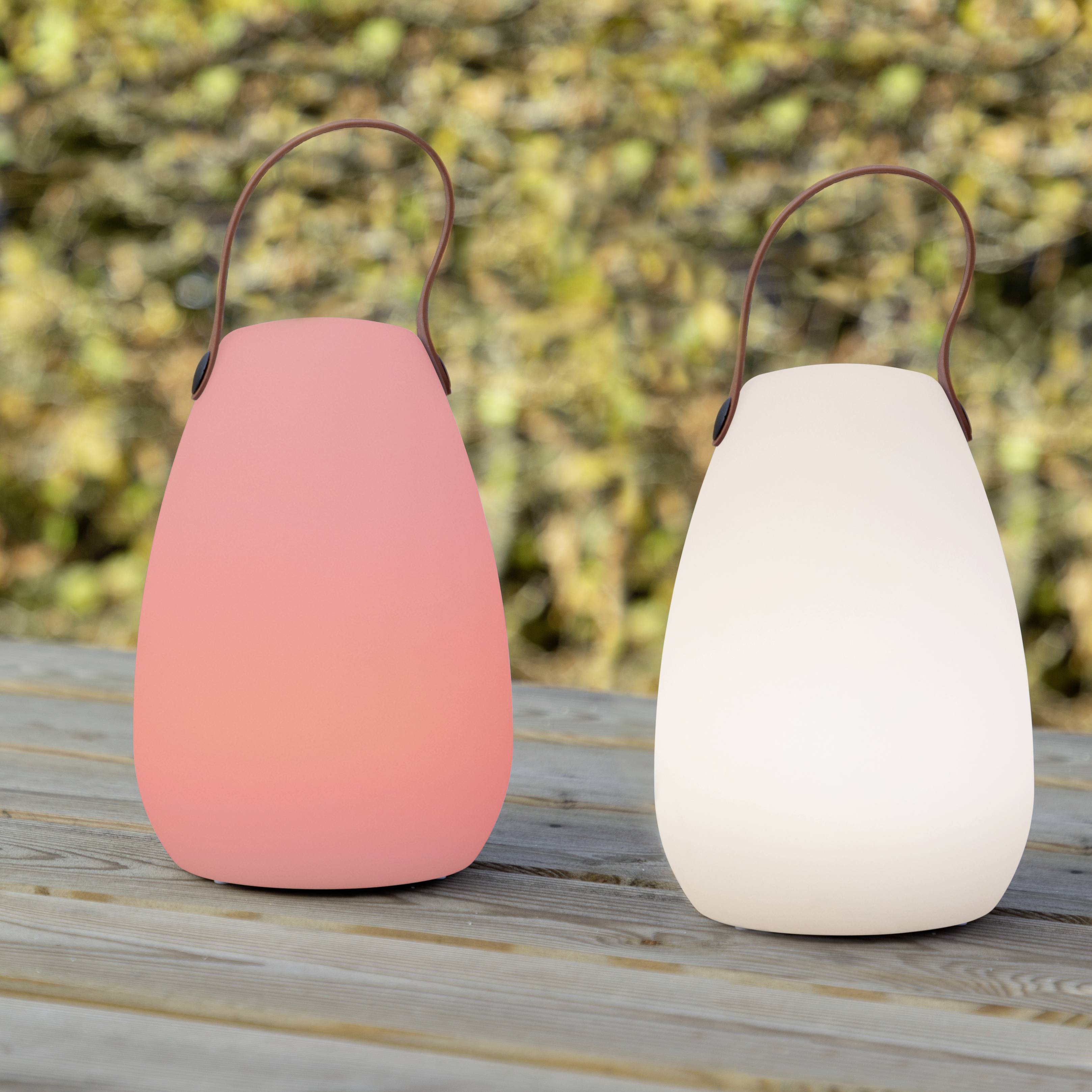 Deux lampes LED portables posées sur une table en bois à l'extérieur, l'une rose et l'autre blanche, munies de poignées. Parfaites pour un éclairage d'ambiance.