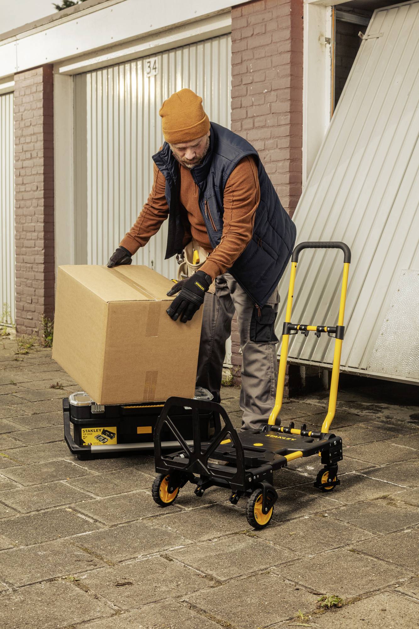 Un homme soulève un grand carton devant une porte de garage. À côté de lui se trouve un chariot jaune sur un chemin pavé.