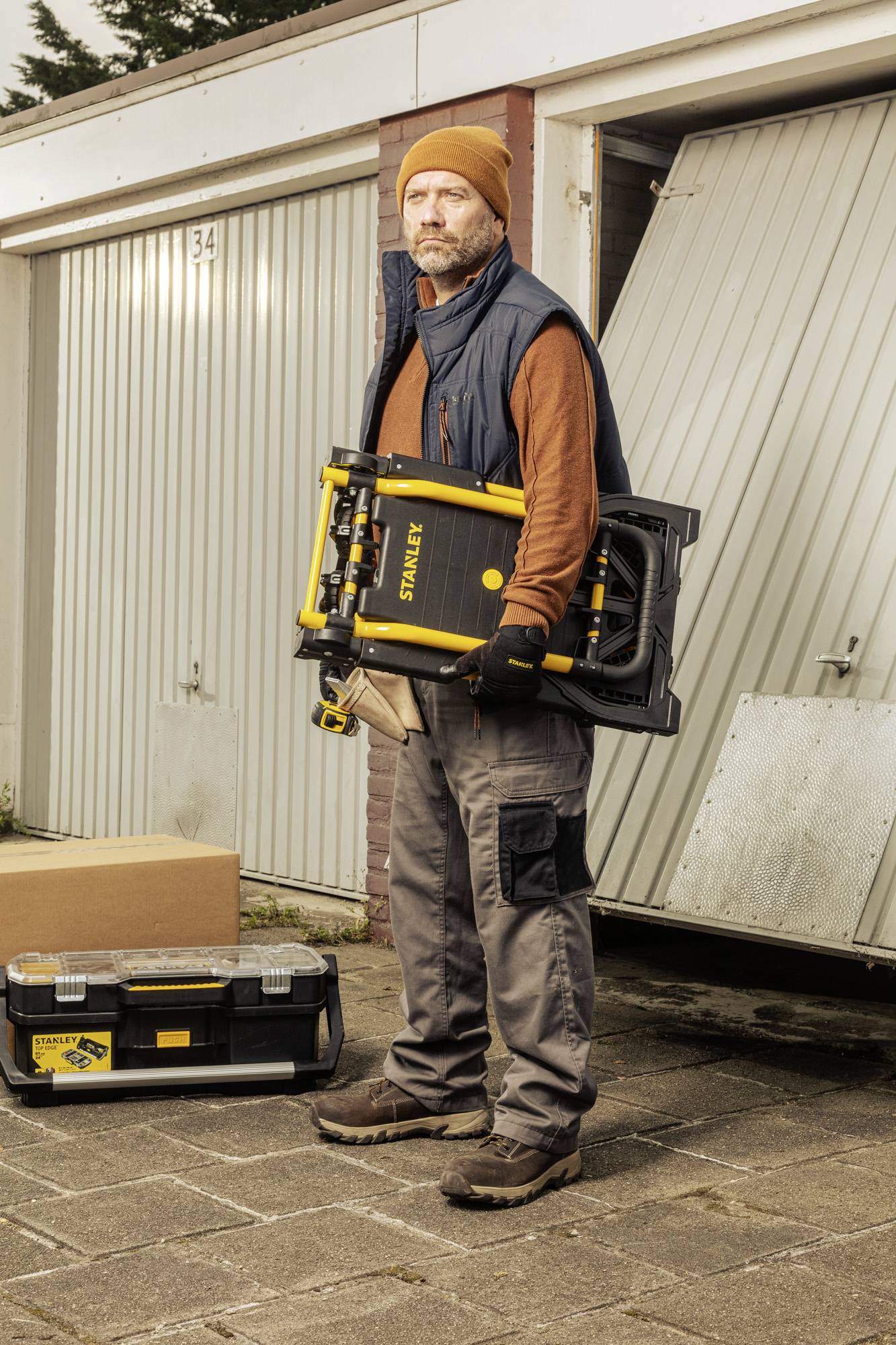 Un homme portant une casquette et des gants se tient devant un garage, tenant un outil jaune, entouré d'une boîte à outils et d'un carton.