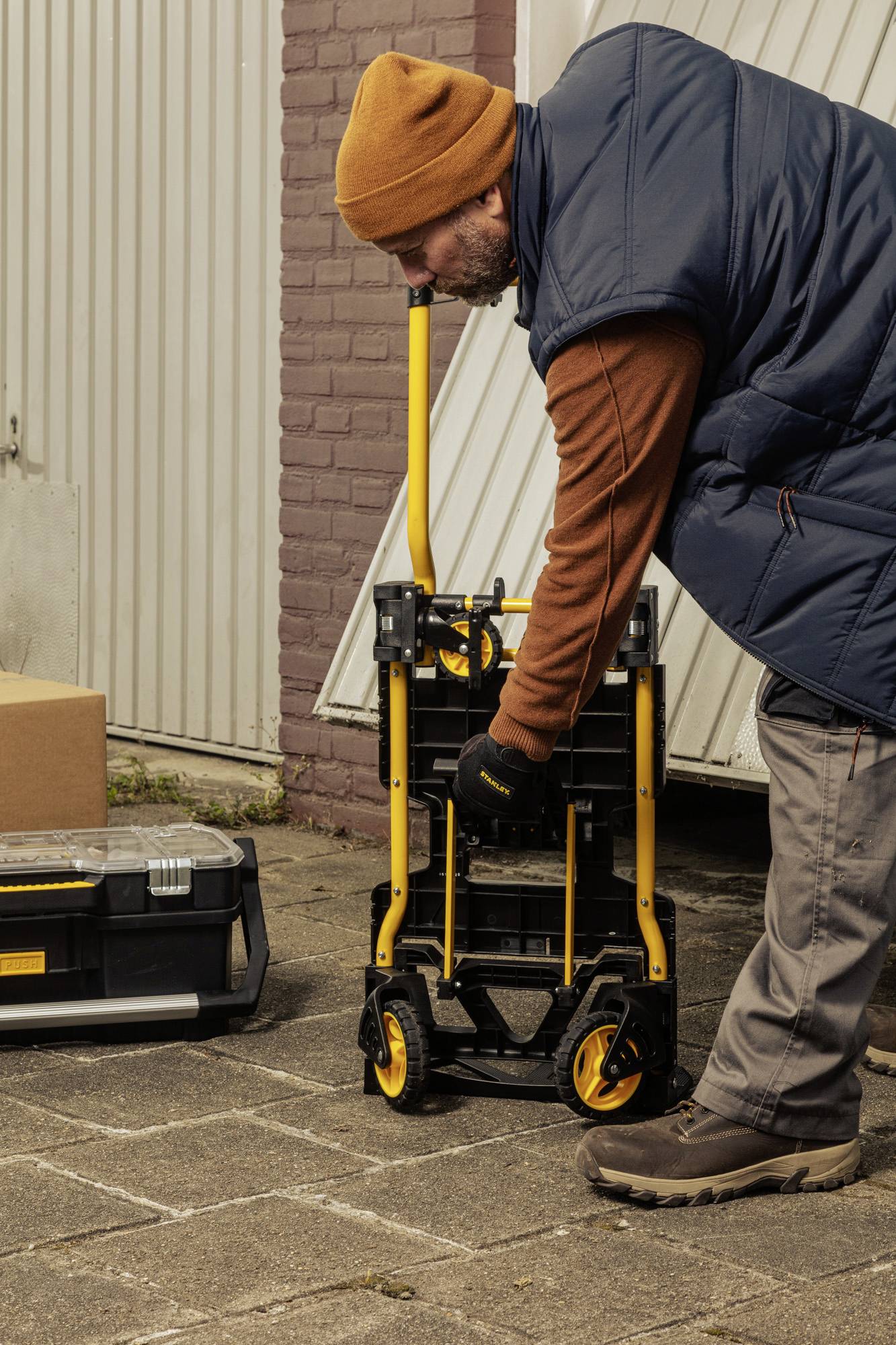 Un homme tire une charrette à bras avec des roues jaunes devant un garage. À côté se trouve une boîte à outils. Il porte une casquette orange et des gants.