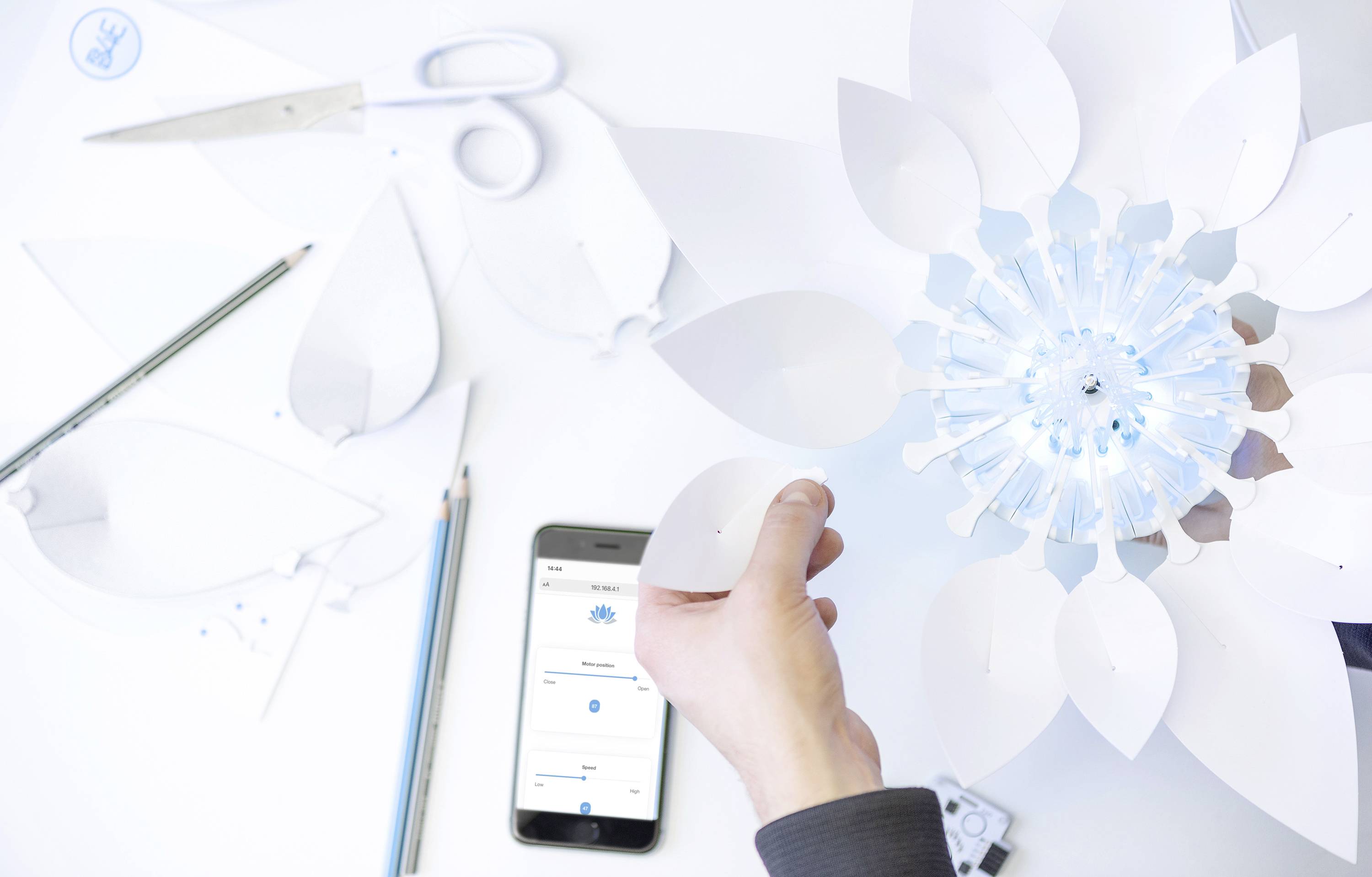 Une personne tient une fleur en papier. Des ciseaux, des crayons et un smartphone avec une application sont posés sur la table. Un travail lumineux et créatif.