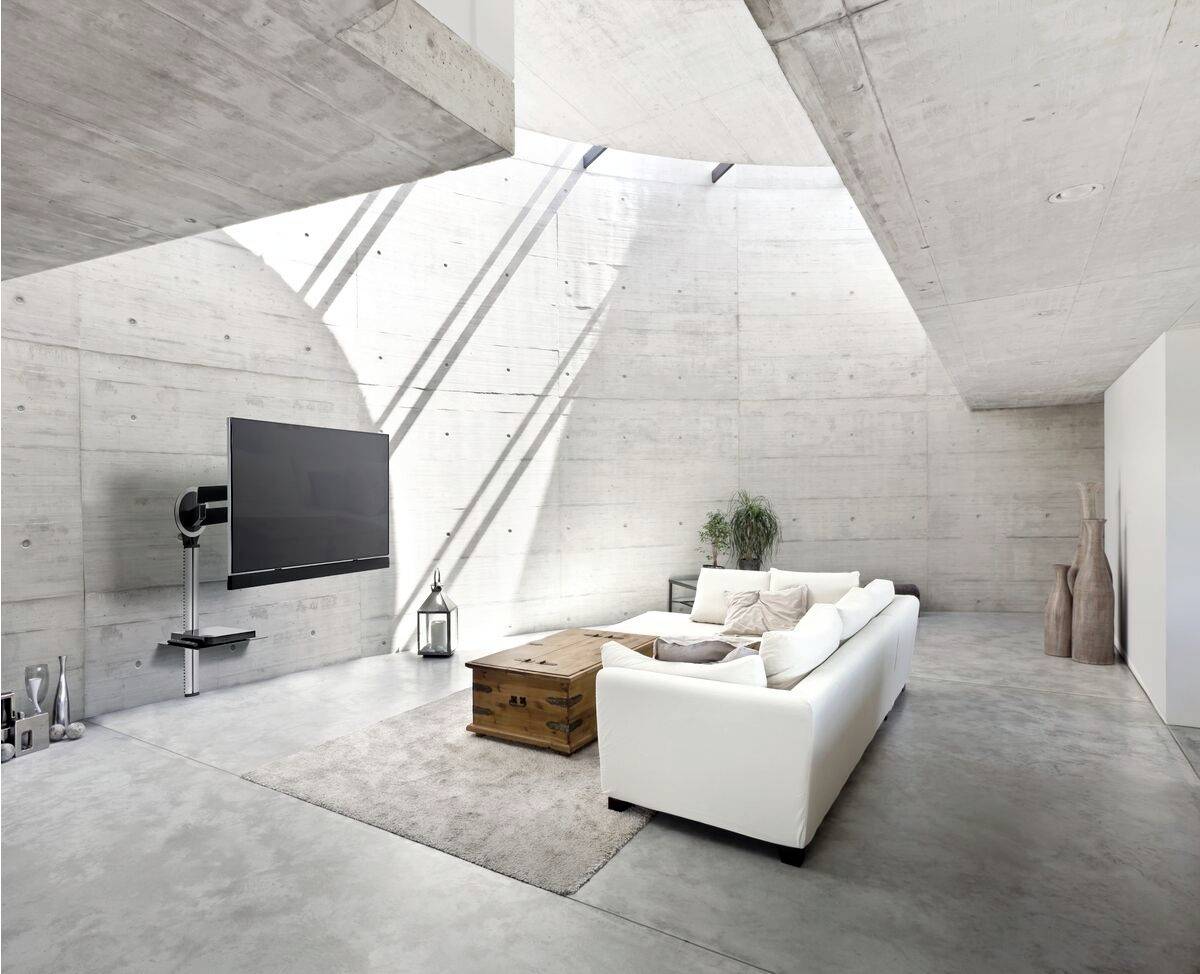 Salon moderne en béton avec un grand canapé, une télévision murale et une décoration minimaliste. Beaucoup de lumière naturelle entre par les lanterneaux.