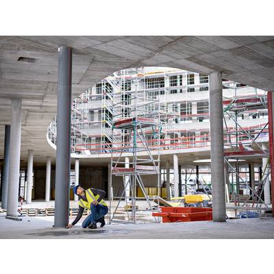 Un ouvrier est à genoux sur un chantier, en train de fixer une poutre métallique. À l'arrière-plan, un système d'échafaudage complexe et un bâtiment inachevé sont visibles.