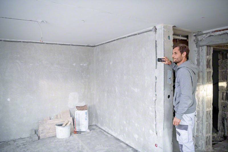 Un homme en tenue de travail mesure un mur dans une pièce inachevée à l'aide d'un télémètre laser. Du matériel de chantier est visible en arrière-plan.