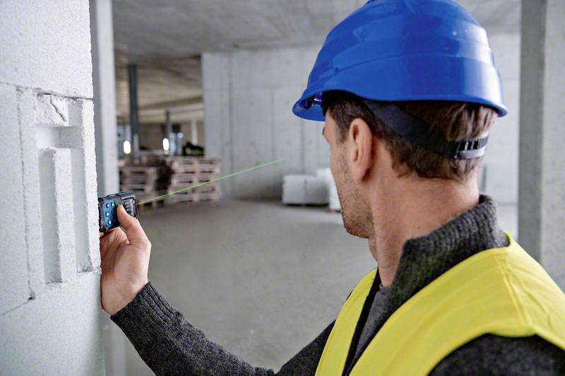 Un ouvrier portant un casque bleu et un gilet jaune utilise un télémètre laser sur un mur en béton dans un chantier de construction.