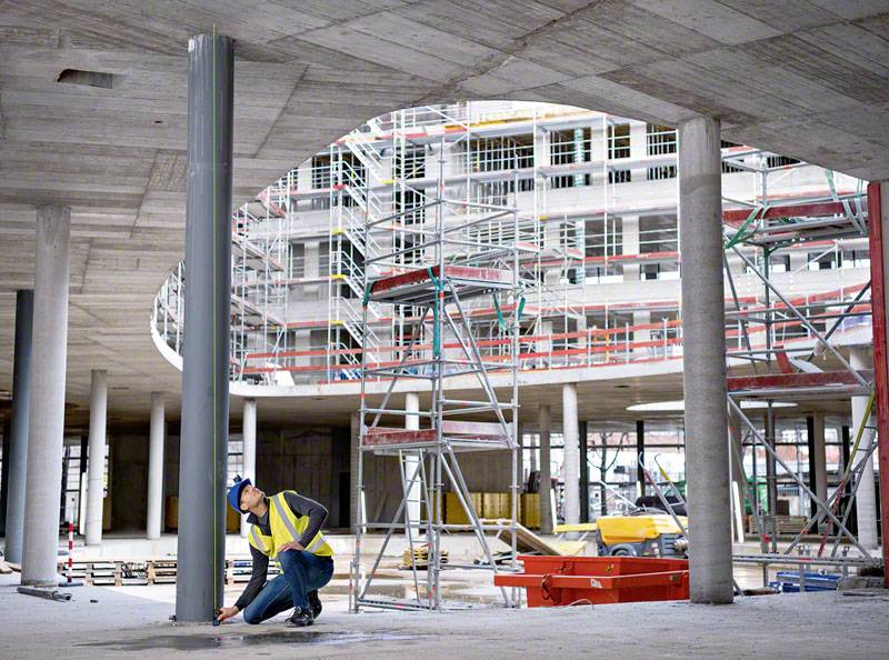 Un ouvrier portant un casque et un gilet de sécurité vérifie un revêtement de sol dans un chantier moderne en construction, entouré d'échafaudages et d'équipements de construction.