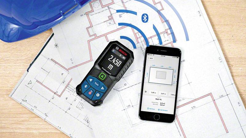 Smartphone et télémètre laser sur des plans de construction, connectés via Bluetooth. Le laser affiche '2,450 m', l'application montre la mesure.