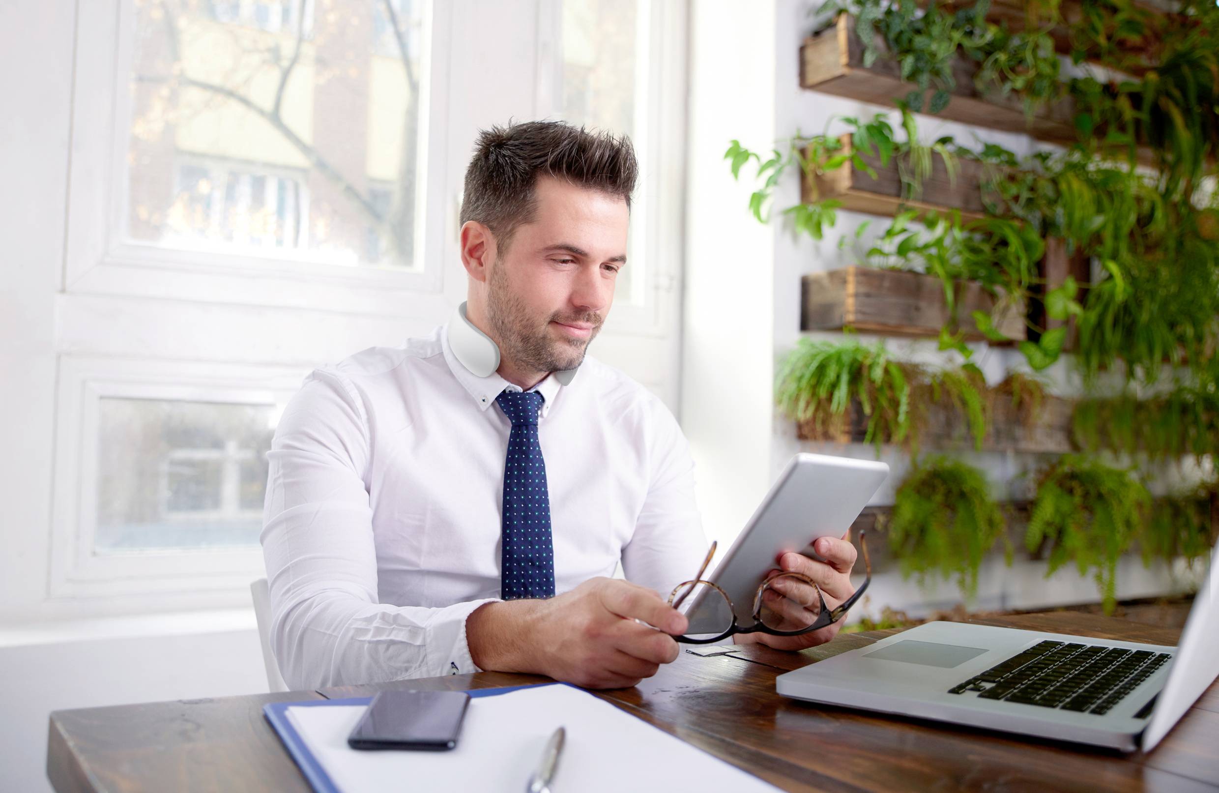 Un homme portant une chemise blanche est assis à un bureau avec un ordinateur portable et une tablette. Des plantes sont disposées sur le mur en arrière-plan.