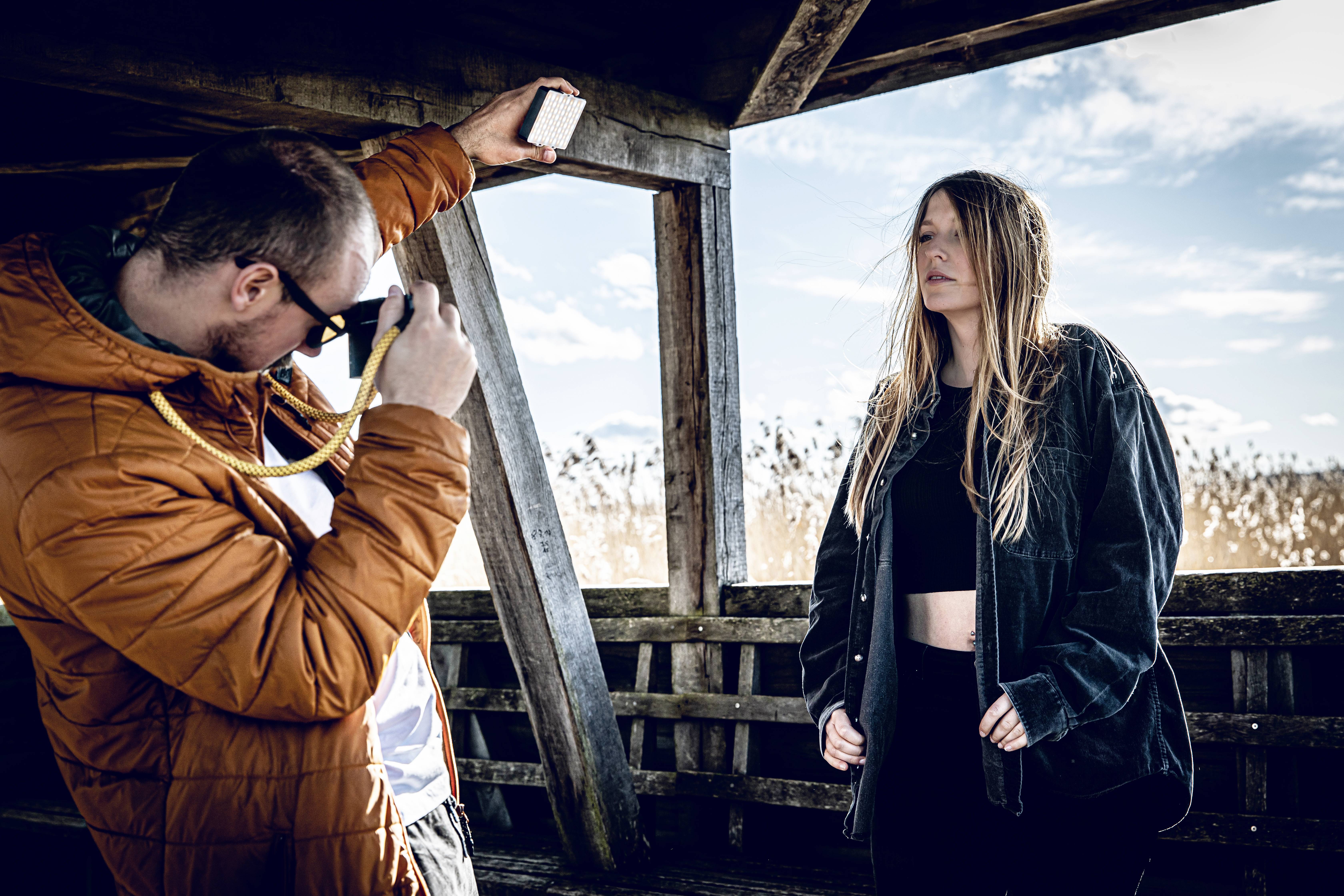 Une personne photographie une autre personne sous un abri en bois. L'environnement semble rural, avec un ciel bleu et des nuages.