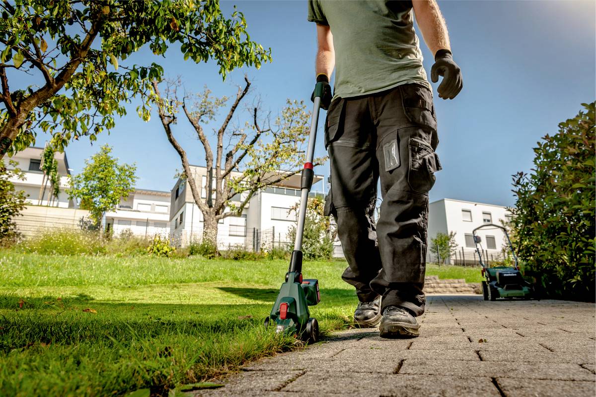 Un homme qui marche le long d'un chemin pavé dans un jardin et tond la pelouse avec un outil de jardinage. Des maisons résidentielles sont visibles en arrière-plan.
