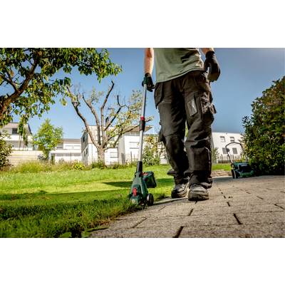 Une personne tond l'herbe le long d'un chemin pavé dans un jardin ensoleillé à l'aide d'un coupe-bordures.