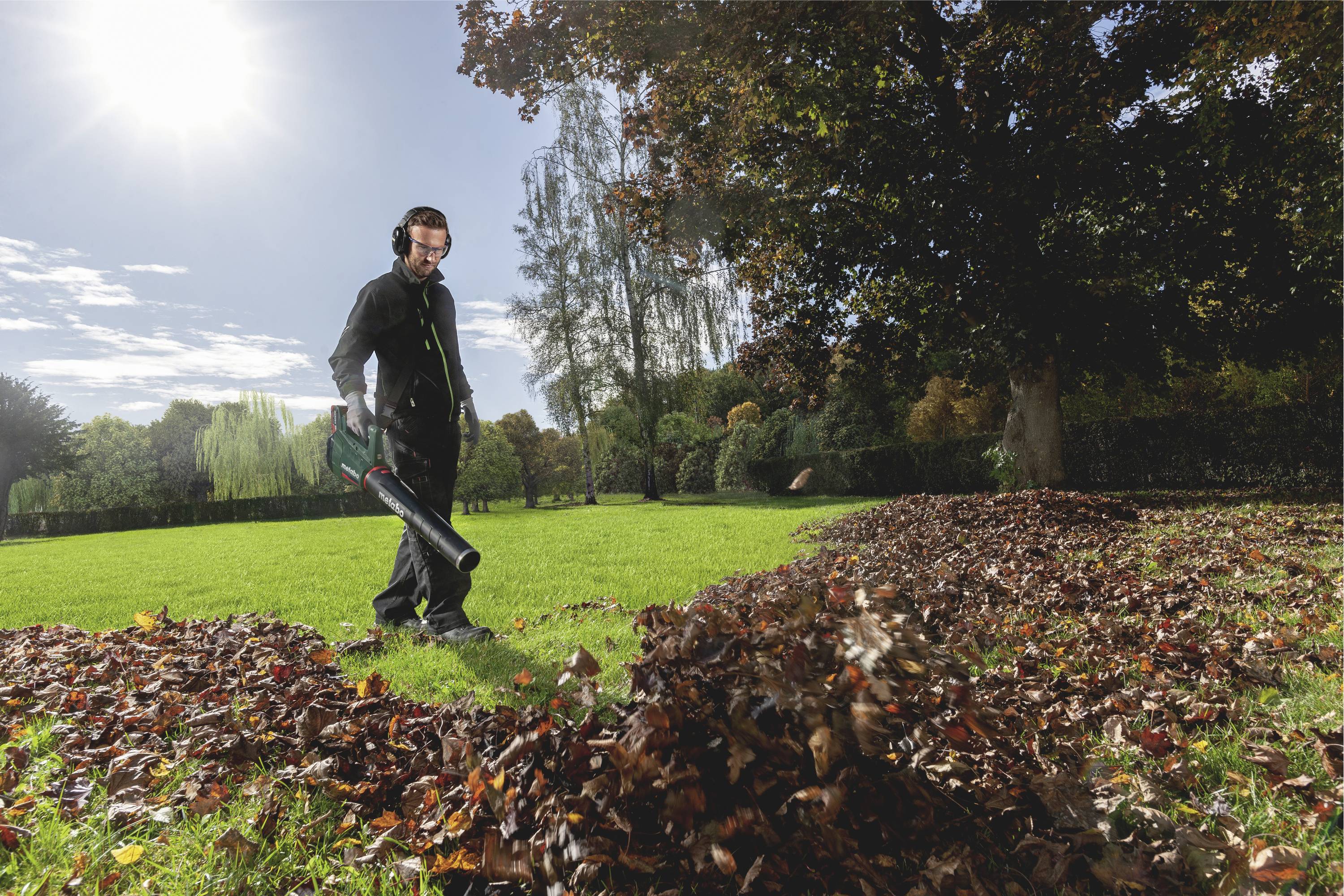 Une personne souffle des feuilles avec un souffleur sur une pelouse, entourée d'arbres automnaux et de tas de feuilles, par une journée ensoleillée.