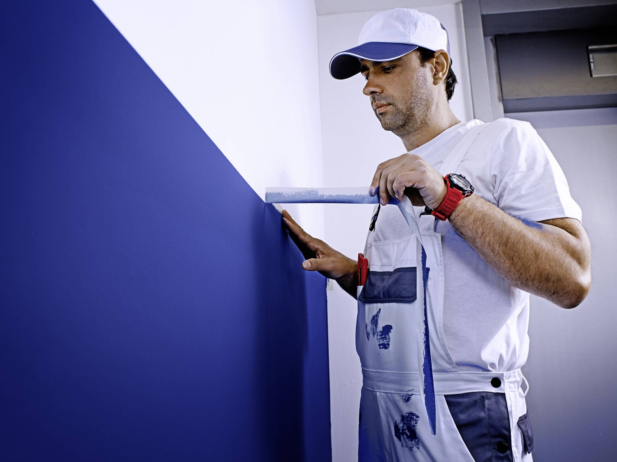 Un homme en combinaison mesure la hauteur du mur bleu avec une règle. Il porte une casquette blanche et une montre.