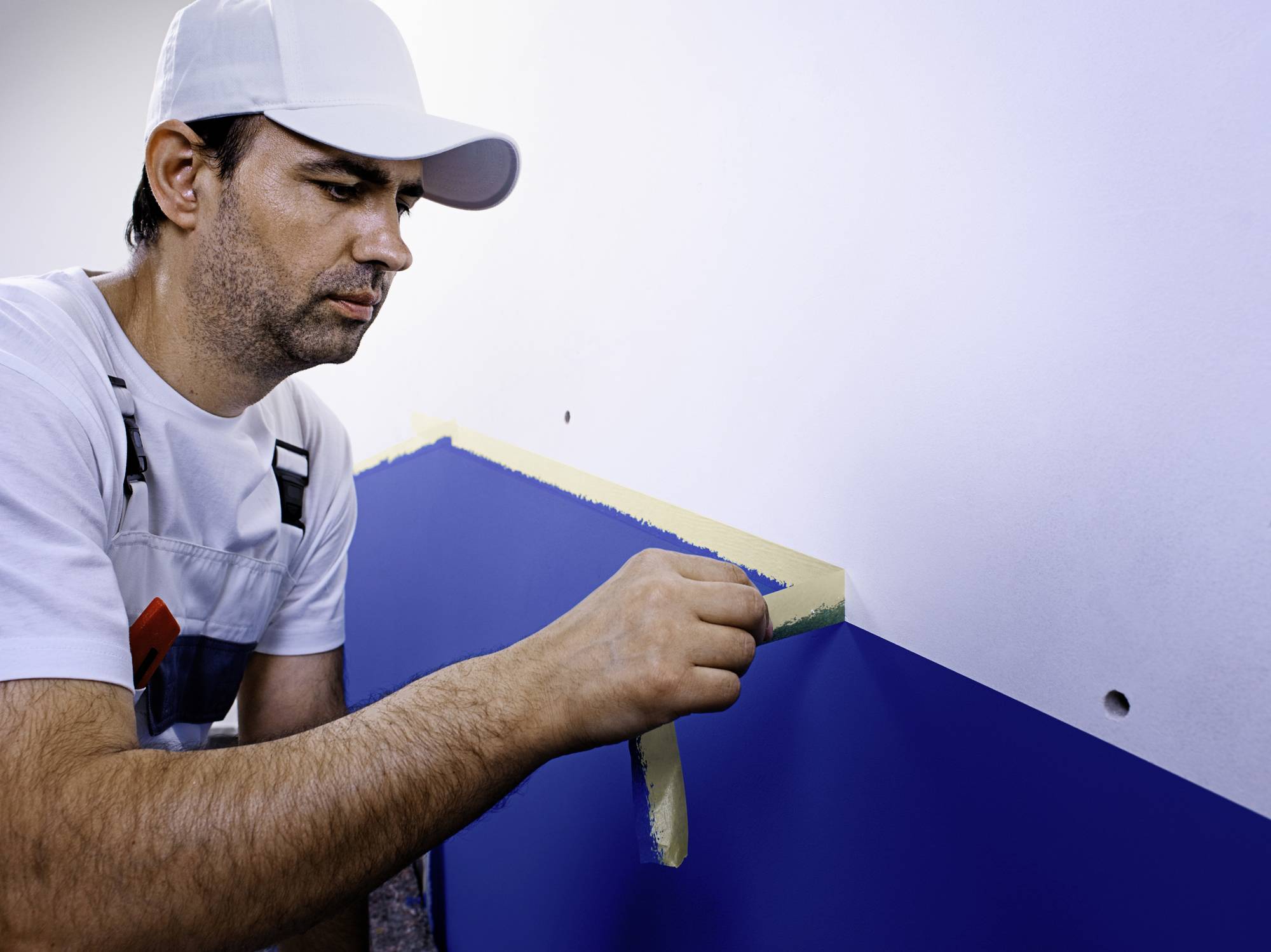 Un peintre en tenue de travail pose du ruban de masquage sur un mur partiellement bleu pour le peindre en deux couleurs.