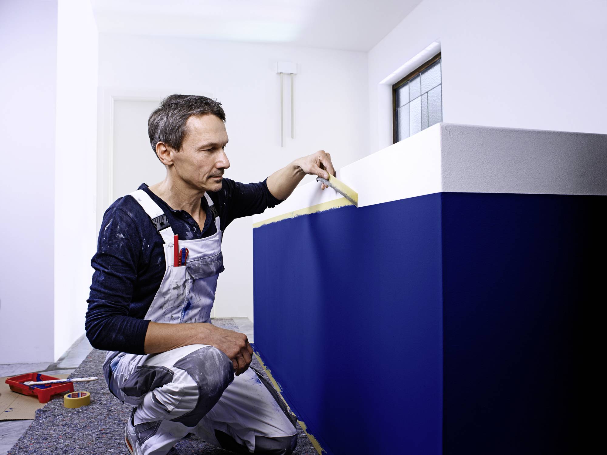 Un peintre en tenue de travail peint un mur en bleu, en se concentrant sur un travail soigné des bords.