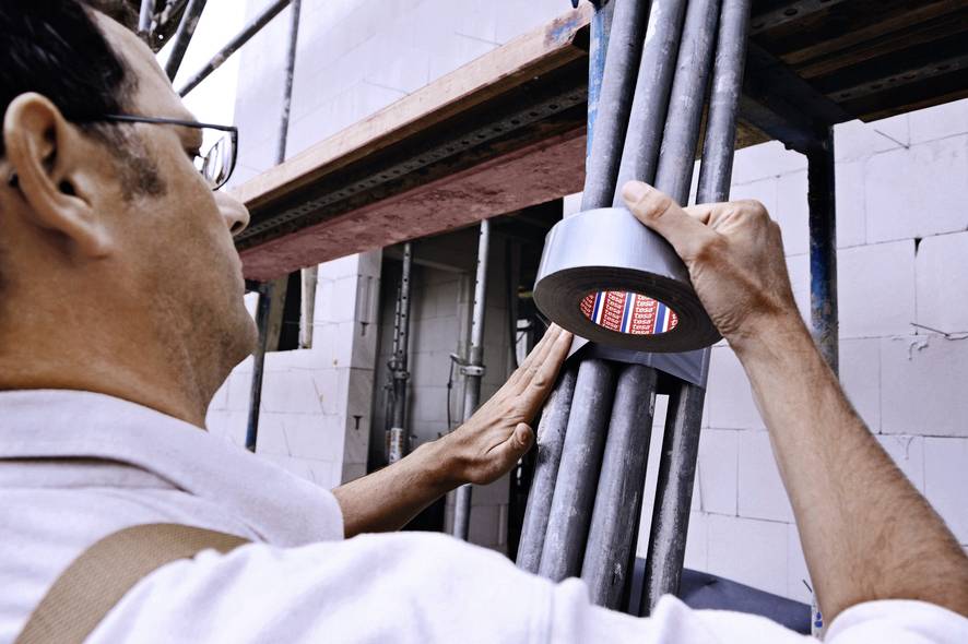 Un homme inspecte des gaines de câbles avec un appareil de contrôle sur un chantier de construction. Il porte une chemise blanche et des lunettes.