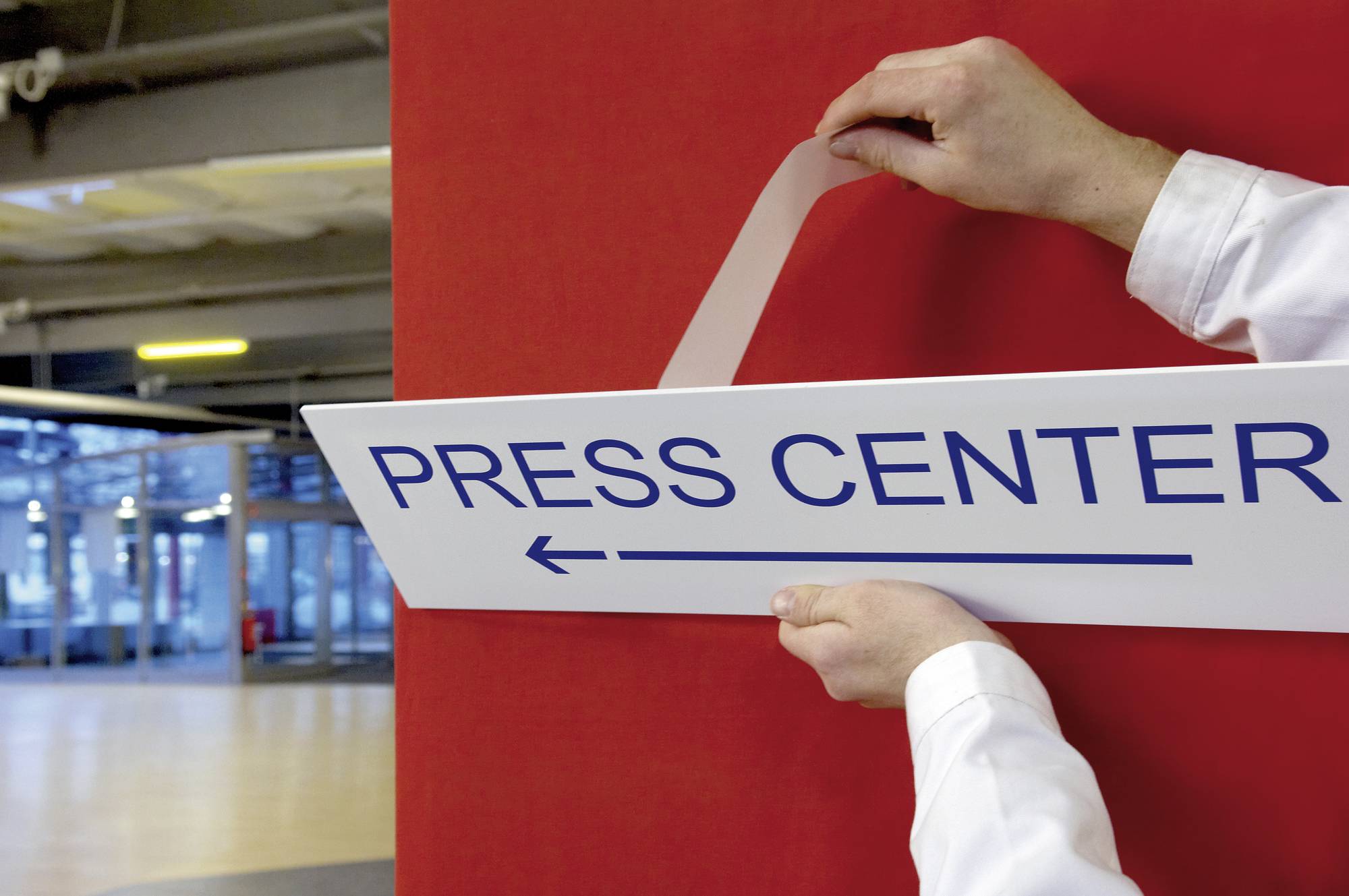 Une personne fixe un panneau portant l'inscription 'PRESS CENTER' sur un mur rouge dans un bâtiment. Une flèche indique une direction vers la gauche.