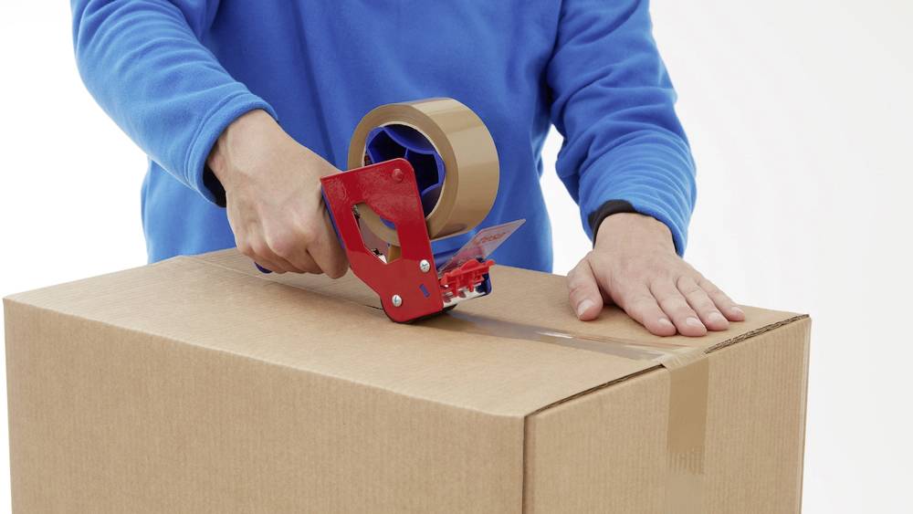 Une personne portant un pull bleu ferme un carton avec du ruban adhésif.