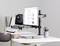 Une personne travaille à un bureau avec un ordinateur portable et un écran, qui sont installés sur des supports réglables. À côté, un porte-stylos et des livres sont disposés.