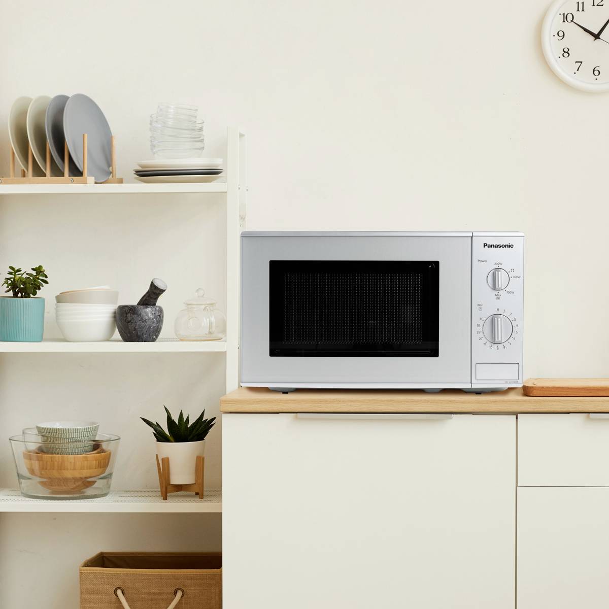 Un micro-ondes est posé sur un plan de travail de cuisine. À gauche se trouvent des étagères avec de la vaisselle et des plantes. À droite, une horloge est visible.