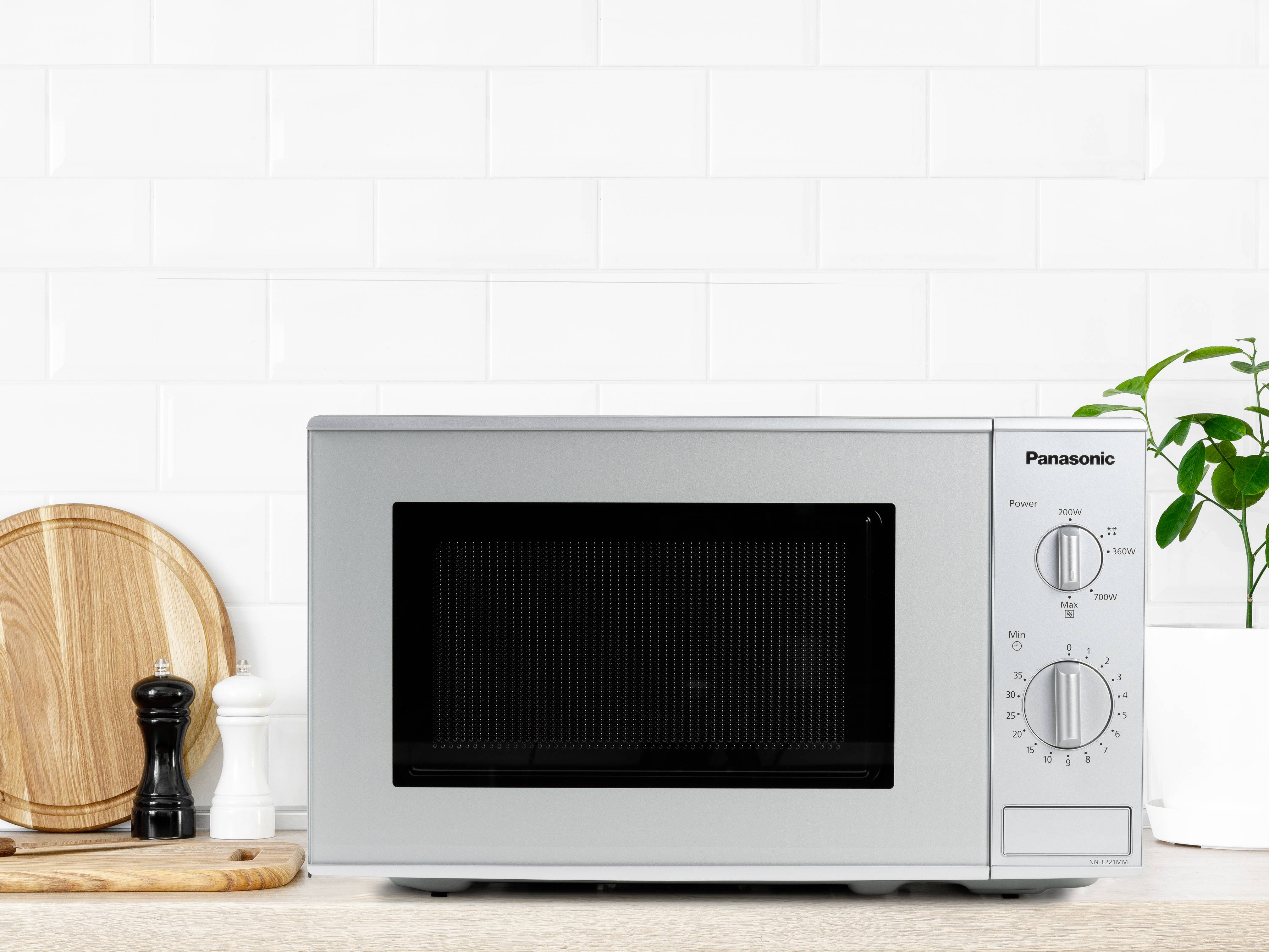 Four micro-ondes sur un comptoir de cuisine, entouré d'une planche à découper en bois et de moulins à épices, devant un mur de carrelage blanc.