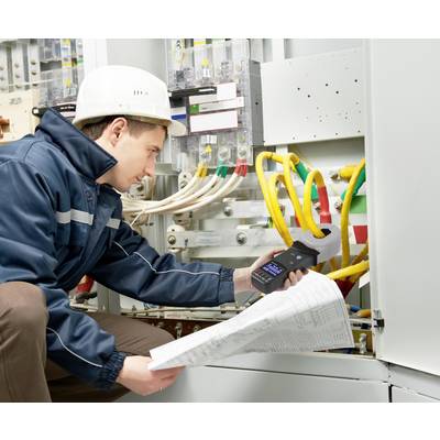 Un technicien portant un casque de protection vérifie les connexions électriques dans un armoire électrique. Il tient un appareil de mesure et des plans de construction à la main.